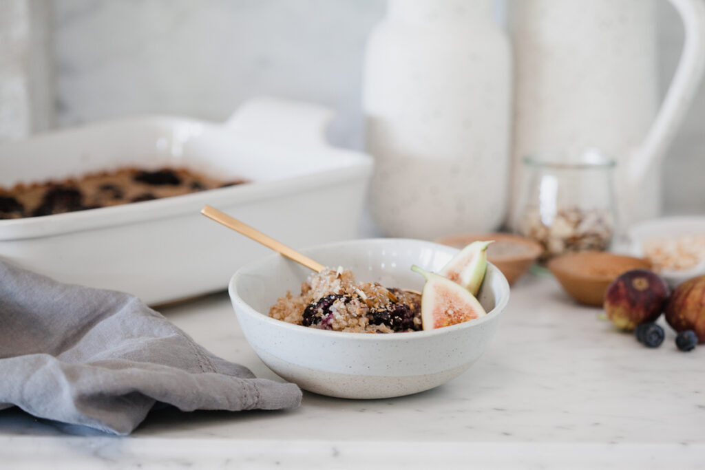 Baked Steel Cut Oatmeal topped with berries and nuts in a white bowl sitting on a kitchen counter next to a casserole dish